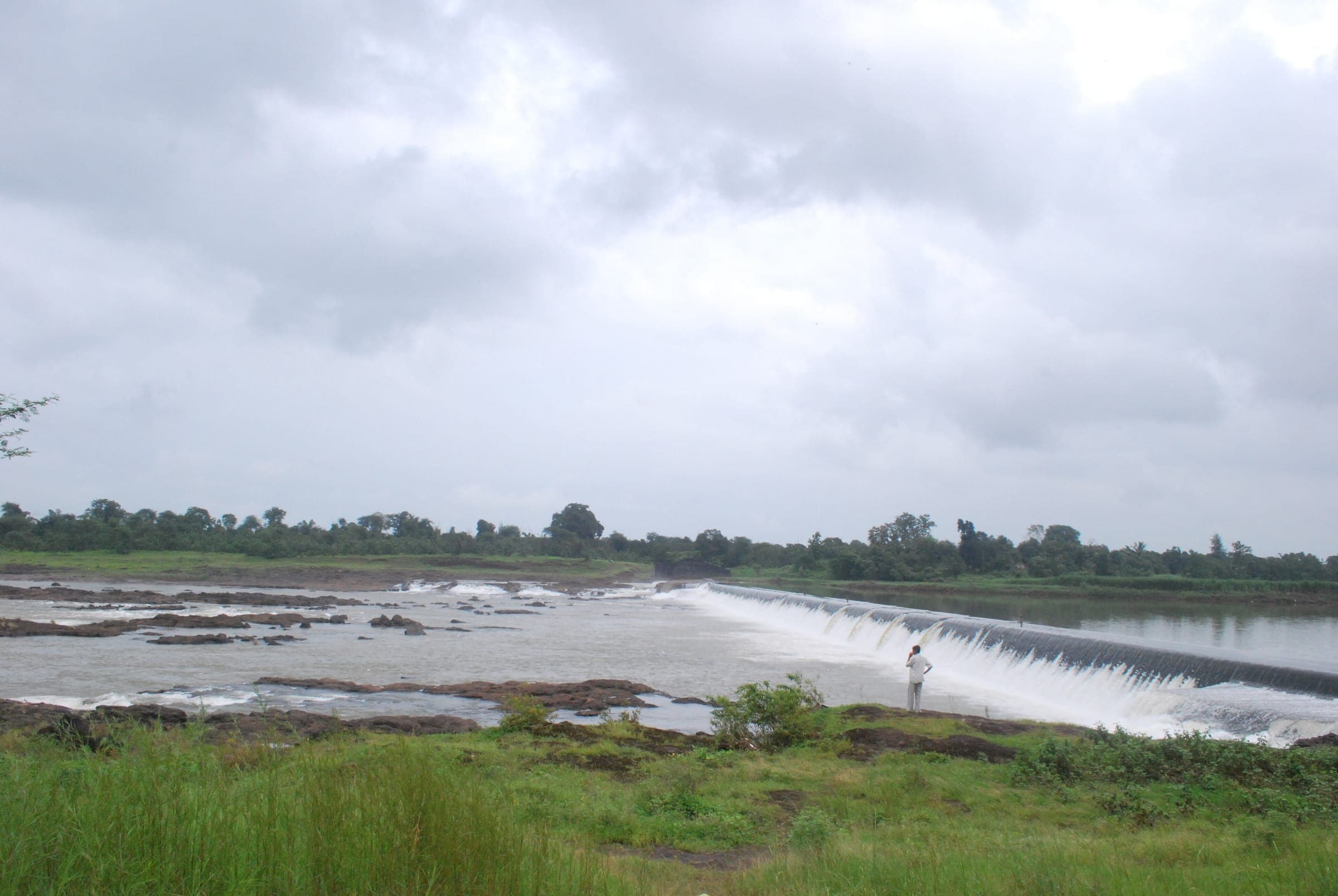 Water flowing over a long check dam across a river under a grey monsoon sky